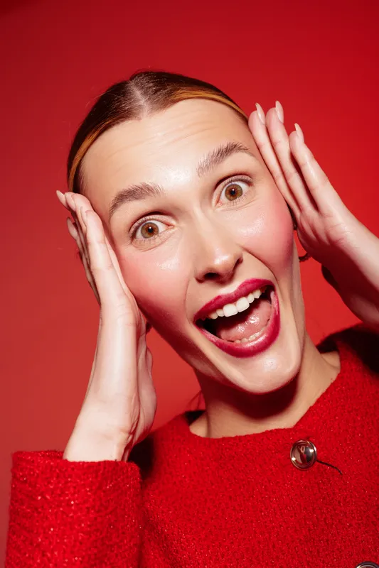 Excited young woman in red sweater with open mouth and hands on face expresses surprise and joy on red background. Concept of emotion, happiness, and positive reaction.