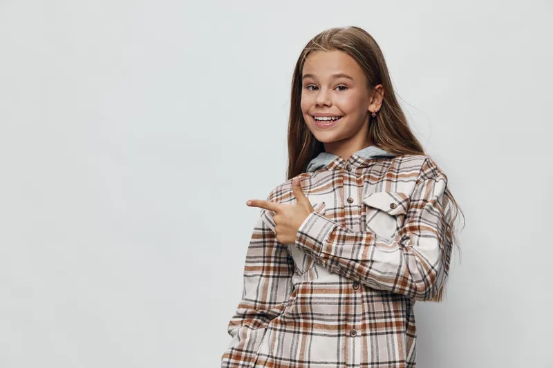 Teenage girl with long brown hair smiling and pointing to side wearing a checkered shirt on light background. Happy young female student with casual style and confident pose.