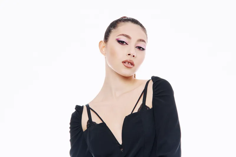 Young woman with elegant makeup and slicked back hair wearing a black fashion outfit poses confidently against white background. Concept of beauty, creative style and glamour.