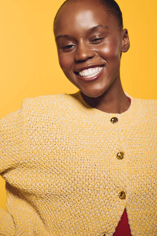 Smiling African woman with dark skin wearing a yellow knitted sweater poses against a yellow background. Concept of happiness, fashion, and natural beauty with bright lighting.