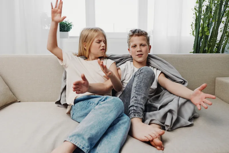 Two children sitting on a couch in casual clothes, showing playful disagreement and expressive gestures in a bright living room with natural light and plants.