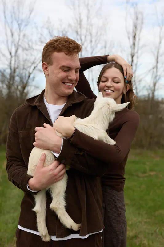 Casual couple outdoors with a playful goat kid, warm smiles, and gentle touch, candid lifestyle moment, soft natural light, relaxed poses, rustic clothing, airy countryside background, wholesome