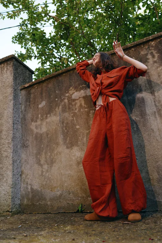 Candid woman lifestyle outdoors in a red jumpsuit standing by a weathered wall, arms raised in a relaxed pose, sunlit scene with lush greenery, natural colors and positive energy
