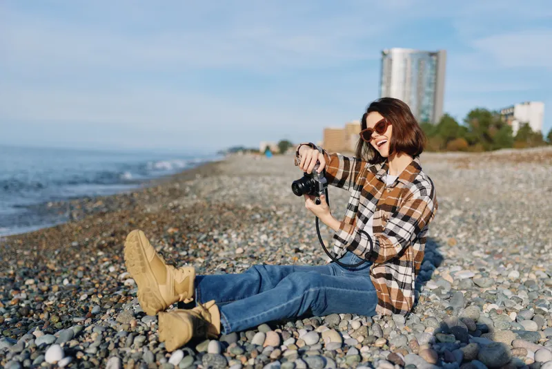 Woman photographer sits on a pebble beach with a camera, wearing a plaid shirt and denim, smiling toward the lens as waves roll in and a distant city skyline shines.
