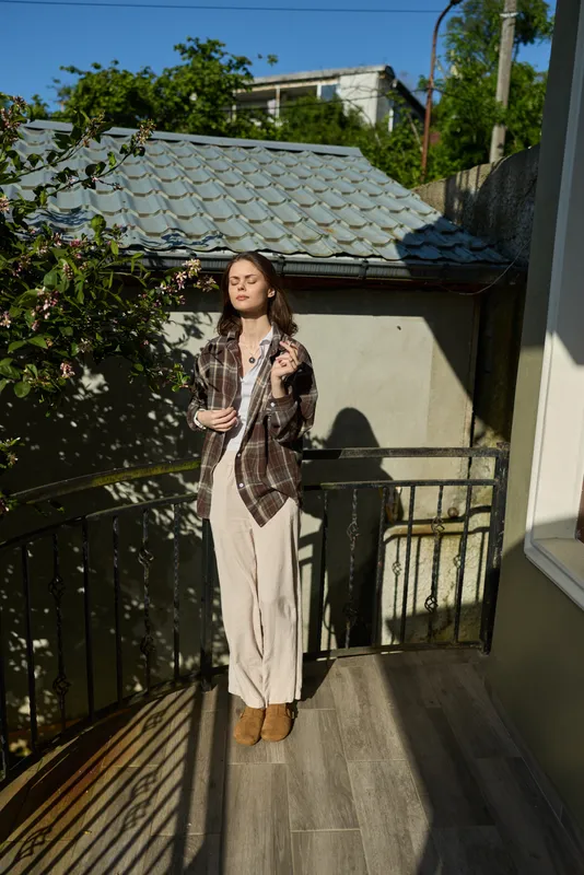 A woman on a sunlit balcony enjoys a relaxed outdoor moment, holding a glass and wearing casual outfit. A calm lifestyle scene that conveys confidence and serenity