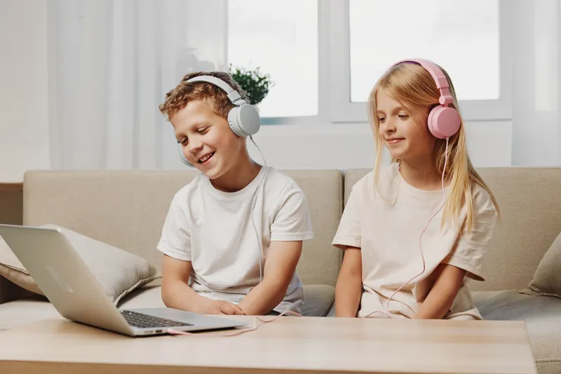 Two smiling children with headphones watch a laptop while sitting on a beige sofa in a bright living room. Concept of online learning, digital entertainment, and childhood technology.