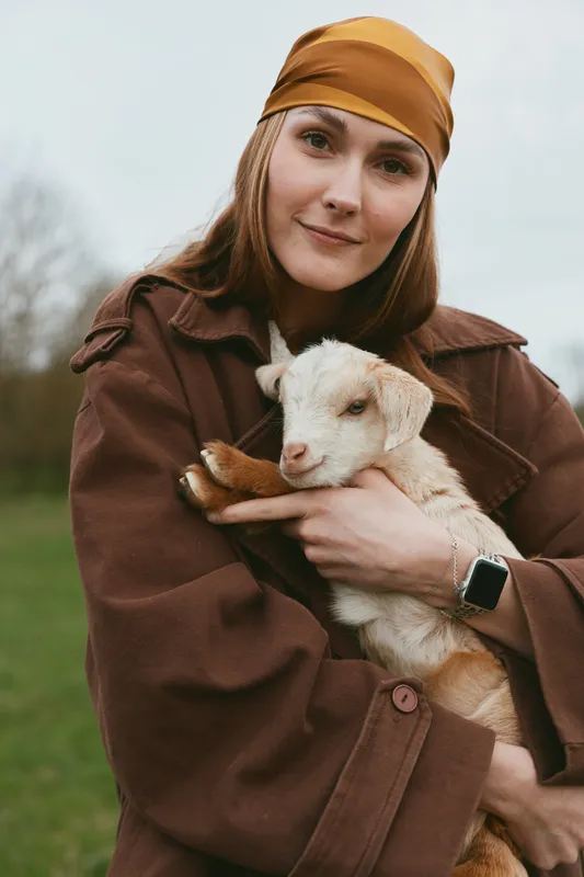 Cheerful lifestyle scene with a woman outdoors, holding a gentle goat kid in her arms, warm coat, soft breeze, natural colors, serene countryside backdrop warmth