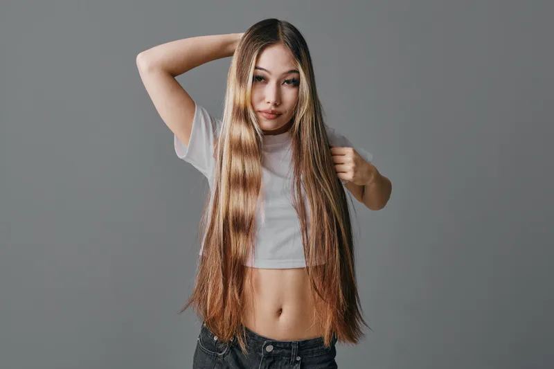 Young woman with long straight hair posing in casual cropped t shirt and jeans against neutral background looking at camera with hand in hair and relaxed expression.