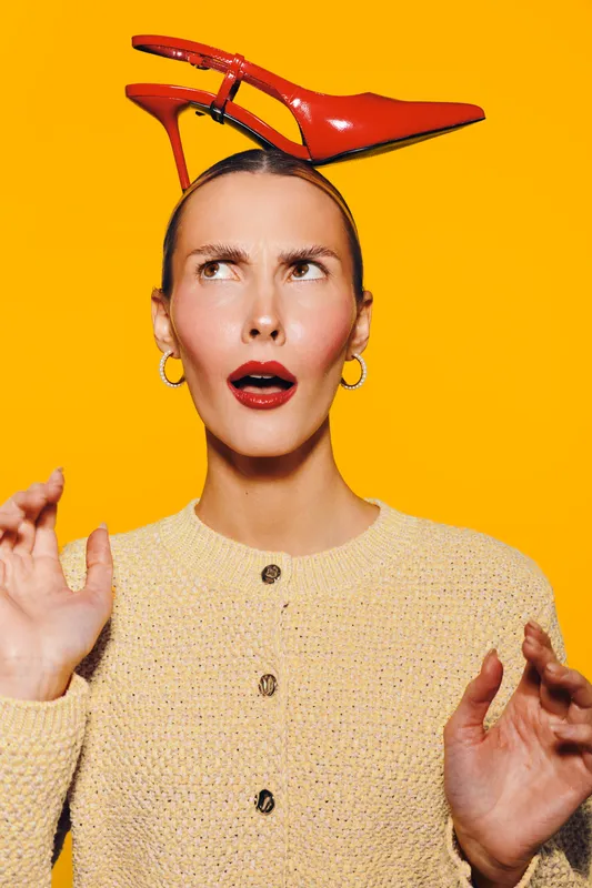 Confused woman with red high heel shoe on head looks surprised, wearing beige sweater and gold hoop earrings. Bright orange background creates vibrant and quirky fashion concept image.