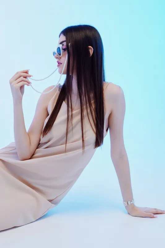 Young woman with long dark hair wearing sunglasses and a beige dress poses seated on a light blue background. Concept of beauty, fashion, and modern style.