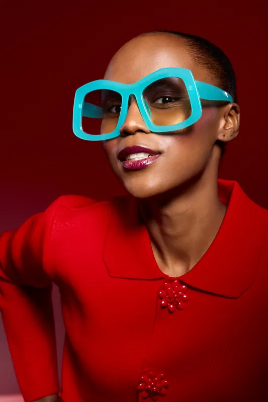 Stylish African American woman with dark skin wearing oversized blue glasses and red outfit poses confidently against red background. Concept of fashion, beauty, and bold accessories.