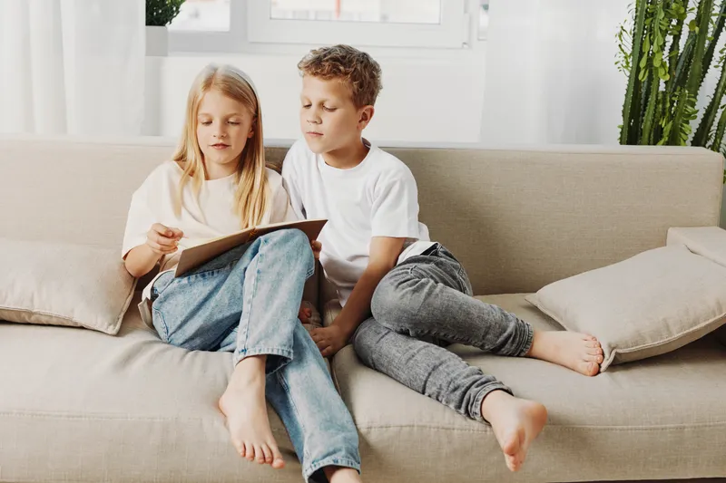 Two children sitting barefoot on a beige sofa reading a book together in a bright living room with natural light and indoor plants.