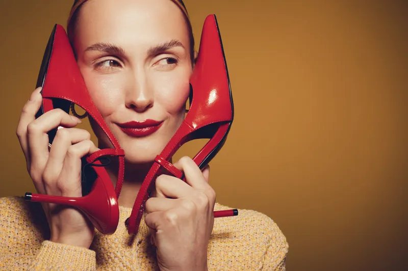 Woman with red lipstick holds red high heel shoes near face, smiling slightly. Warm studio lighting highlights her clear skin and beige sweater. Concept of fashion, style, and beauty.