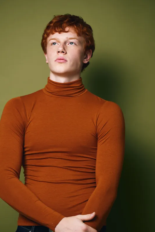 Young man with red hair wearing brown turtleneck sweater standing against green background looking confident with crossed arms and serious facial expression in studio portrait.