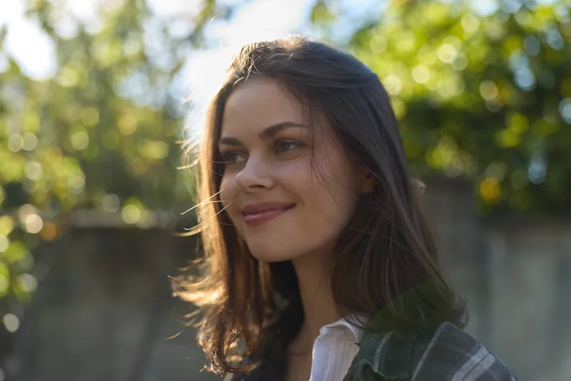 Young woman with long dark hair smiles softly outdoors in warm natural light among green trees, conveying calm confidence and a relaxed lifestyle