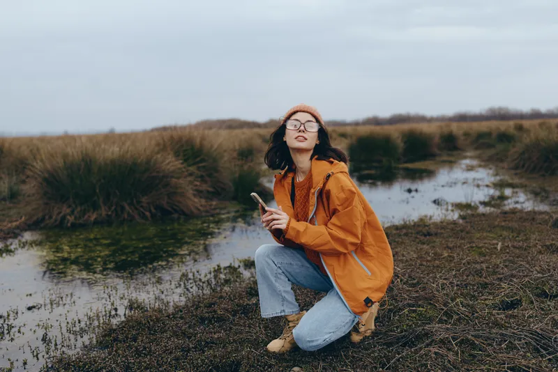 Woman lifestyle outdoors by river in autumn, sitting on grassy bank and reading a book, orange jacket and glasses, calm nature background, soft natural light, weekend leisure