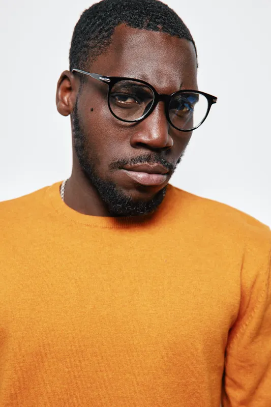 African American man with dark skin wearing glasses and orange sweater looking serious isolated on white background. Portrait, fashion, style and confident expression.
