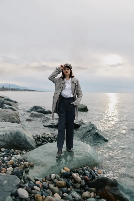 A young woman wearing a trench coat and hat stands on rocky shore near calm sea at sunset. The cloudy sky and peaceful water create a serene coastal atmosphere with fashion style.