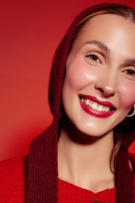 Smiling woman with red lipstick and pearl earring wears red hooded sweater against red background. Close-up portrait with natural skin glow and joyful expression. Concept of beauty and fashion.