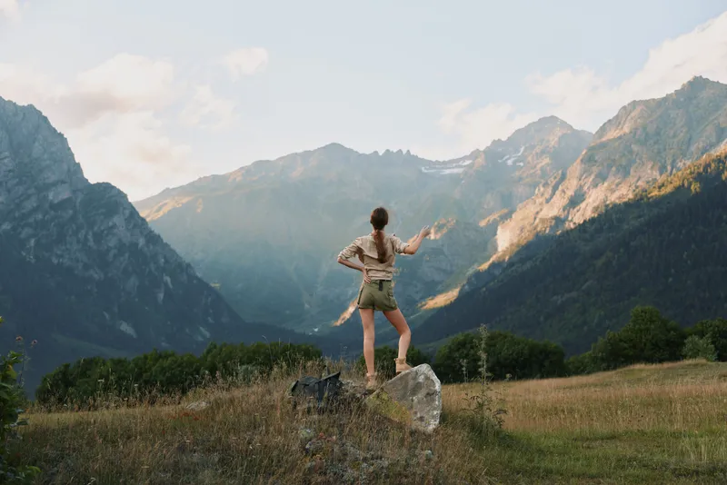 Young woman with long hair stands on a rock in a mountain valley enjoying the scenic view with open arms and casual summer outfit.