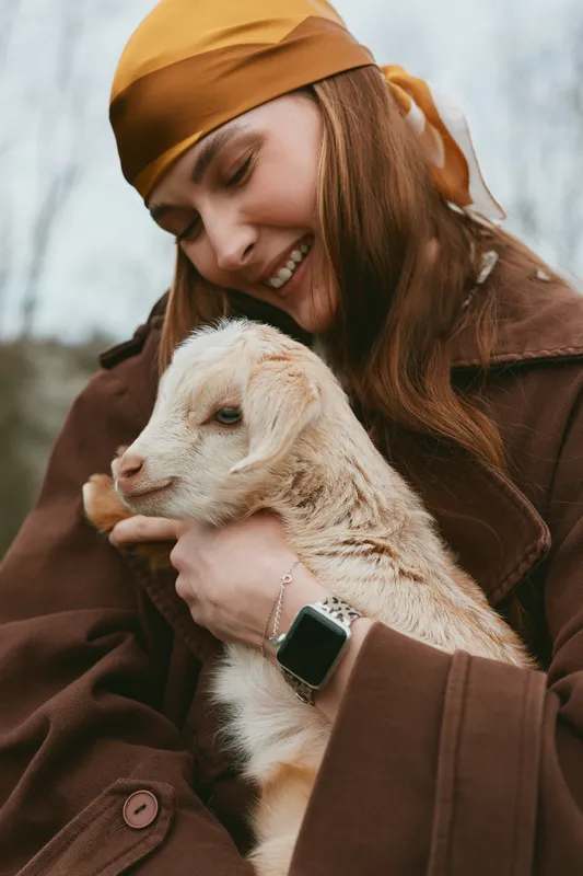 Joyful woman in autumn coat holds a young goat kid close, sharing affectionate moment outdoors, warm scarf, smile, gentle interaction, rustic mood, lifestyle scene, candid portrait, natural light,