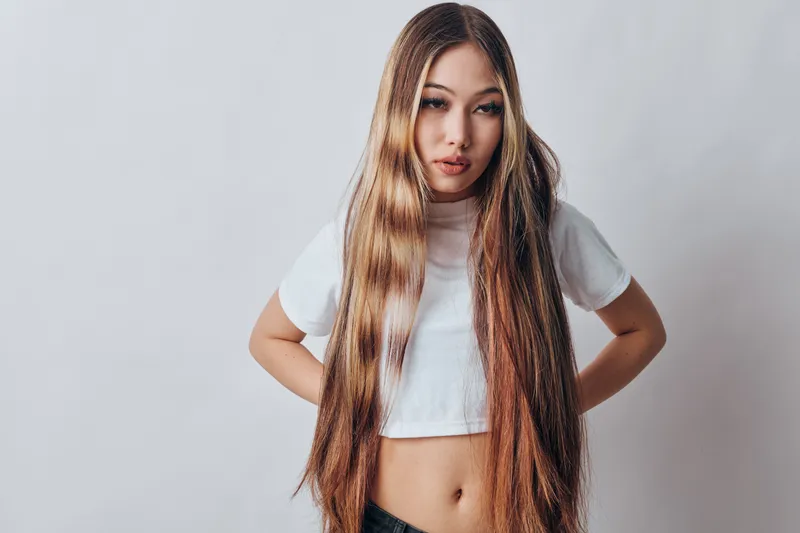 Young woman with long straight hair wearing white crop top and black pants standing with hands behind back on gray background. Beauty, fashion, casual style and confident pose.