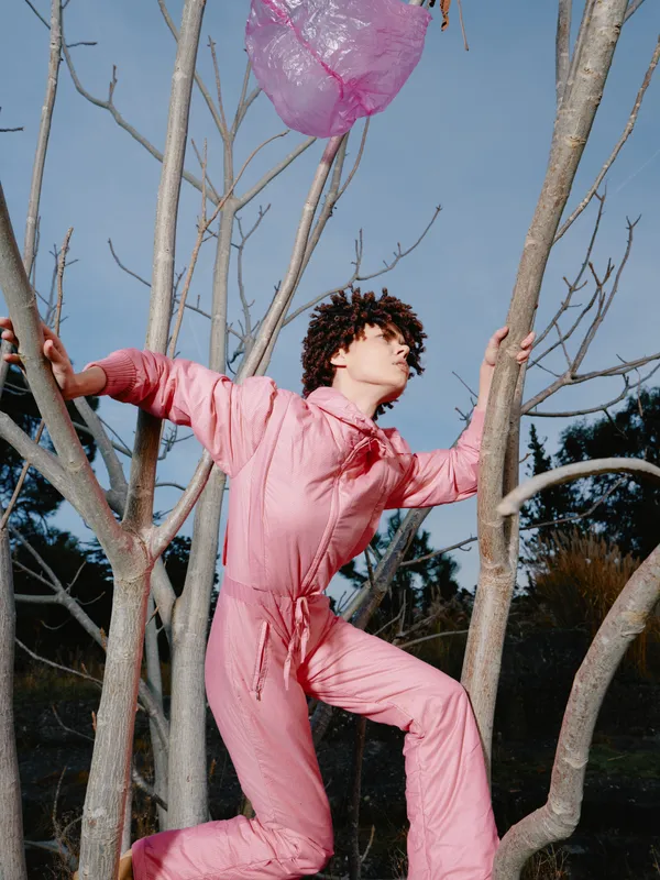 Child climber in a pink jumpsuit among bare tree branches outdoors, reaching for a pink balloon against a blue sky, a playful scene of exploration, nature, and lighthearted adventure