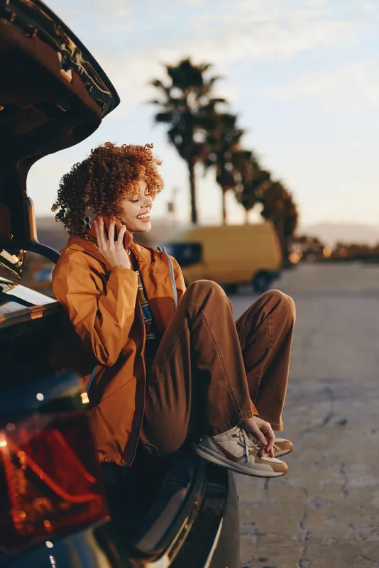 Woman lifestyle outdoors, sitting by open car trunk at sunset, curly hair, phone near ear, relaxed posture, warm golden light, soft street background