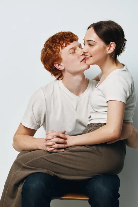 Couple sitting and embracing on chair, young man with red hair and woman with dark hair wearing casual clothes, showing affection and closeness in intimate moment.