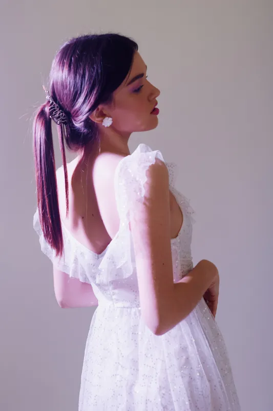 Young woman in a white lace dress poses in profile with closed eyes and pink hair tied back. Soft lighting highlights her beauty and fashion style in a studio setting.