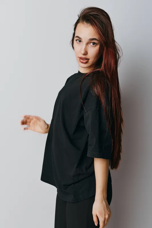 Young woman with long brown hair wearing black oversized t shirt and pants posing with serious expression on gray background in studio. Fashion, beauty and casual style portrait.