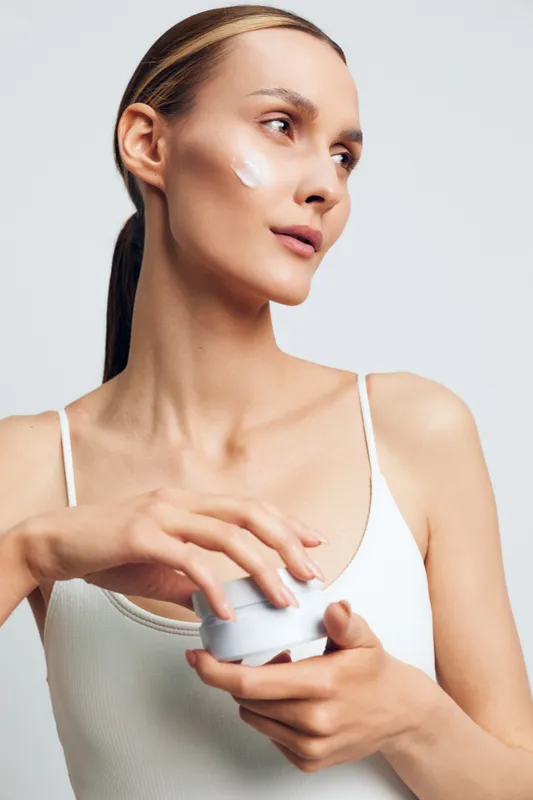 Young woman with clear skin applies moisturizing cream on face holds container looks away wears white tank top isolated on light background skincare beauty concept.