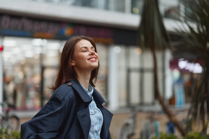 Smiling woman walks along a city street in denim jacket and navy coat with sunny day, urban portrait with blurred background, relaxed casual fashionable mood with poise.