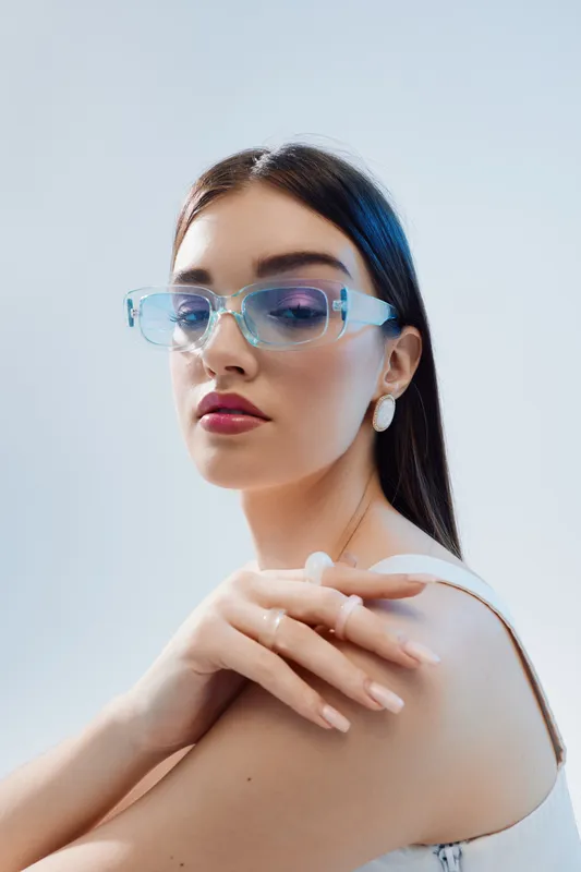Young woman with long dark hair wearing transparent glasses and white earrings poses with crossed hands on shoulder. Studio shot on light background. Concept of beauty fashion and elegance.