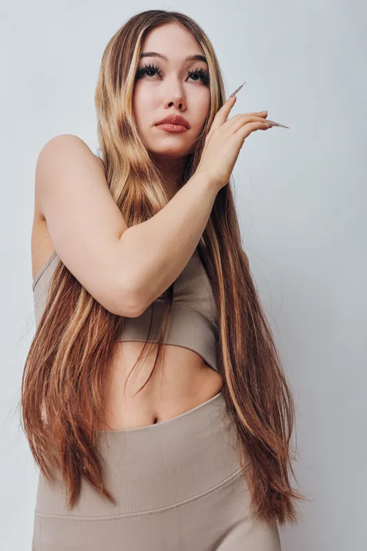 Young woman with long brown hair posing in beige sportswear, fashion style, natural makeup, looking thoughtfully on light background, healthy skin and fitness appearance.