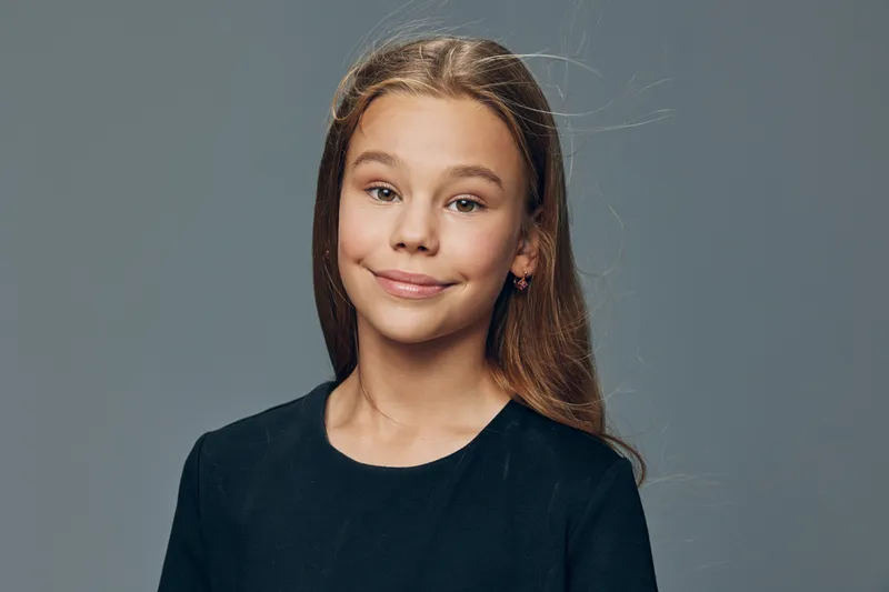 Girl portrait with long hair wearing black shirt looking at camera with slight smile on gray background in studio. Child with natural expression and casual style for kids photography.