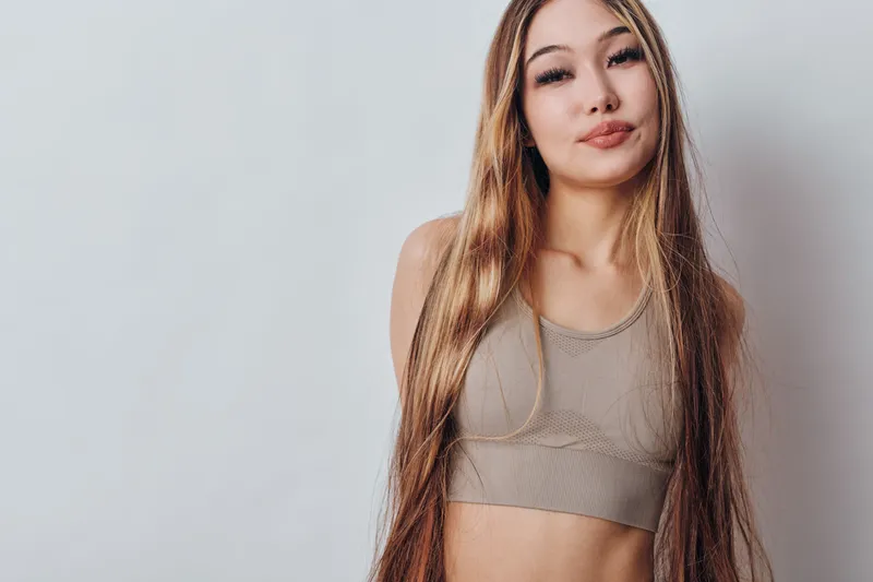 Young woman with long hair wearing a sleeveless beige top stands against plain light background, looking confidently at camera with relaxed expression and natural makeup.
