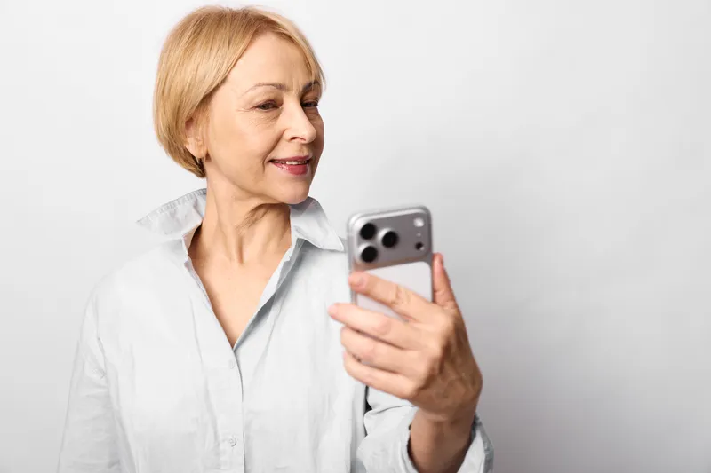 Elderly woman smartphone communication technology texting social portrait smile closeup, she holds a smartphone and smiles softly. Studio closeup on neutral background, digital messaging concept.