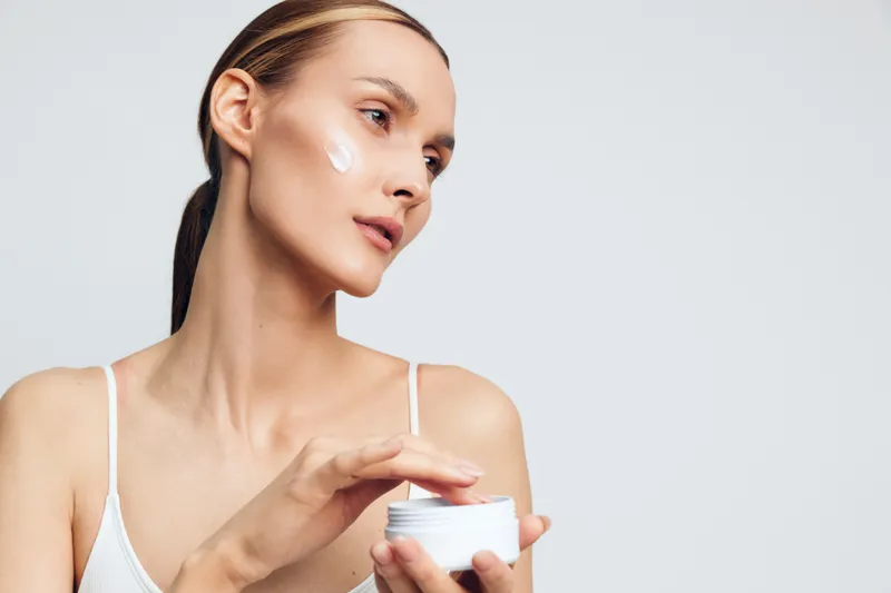 Young woman with clear skin applying white face cream on cheek, wearing white tank top. Concept of skincare, beauty routine, and self-care with soft lighting on light background.