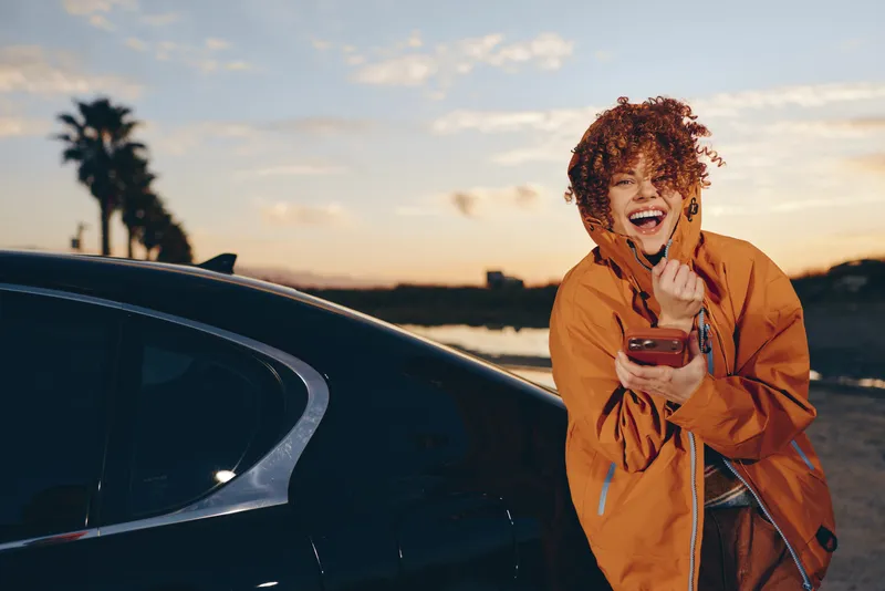 Woman lifestyle near a car at sunset, outdoors, Smiling woman in orange coat holds a phone, gesturing by the road under golden sky, casual travel portrait for getaway