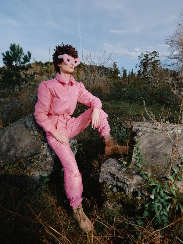 Person wearing a pink jumpsuit and mask poses on rocks in a natural setting, bold fashion statement, dramatic outdoor portrait under a clear sky