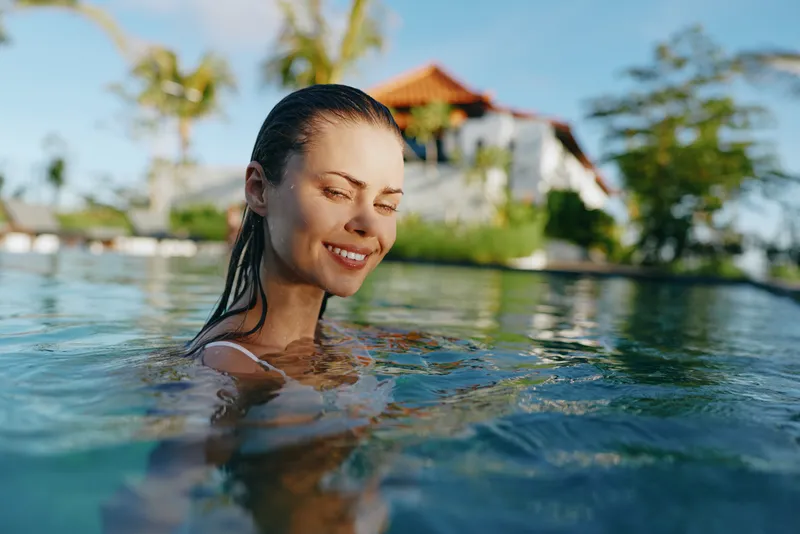 Happy relaxed woman enjoys a refreshing swim in a sunlit tropical pool, expressing serenity and leisure, outdoors at a resort, calm smile amid clear water and bright scenery.