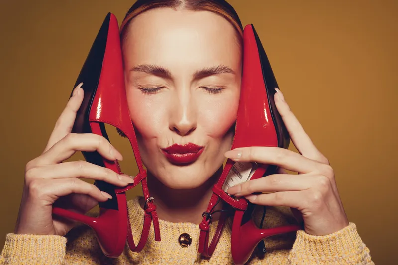 Young woman with closed eyes holds red high heel shoes near face, wearing yellow sweater. Concept of fashion, style, beauty, and footwear with warm studio lighting on brown background.