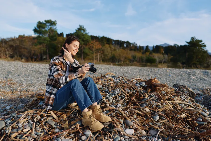 Woman, photographer, camera, outdoor, autumn, nature, beach, casual, lifestyle, hobby, season, scenery, shore, pebbles, forest, trees at calm seaside setting with scenic light and candid mood for