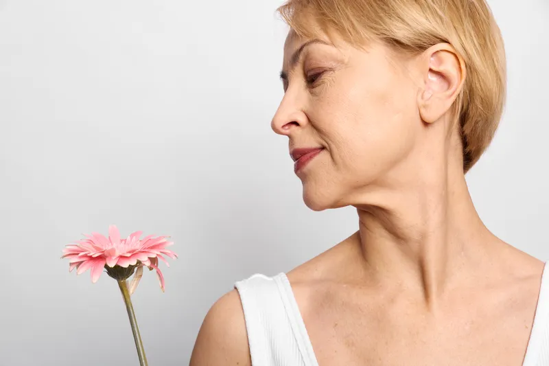 Calm blonde woman in profile holding a pink flower, eyes closed, soft skincare and beauty concept, studio closeup against white background, minimalist wellness mood.