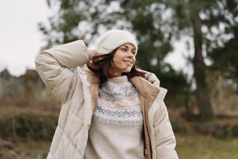 Woman in a warm beige puffer coat and knit sweater outdoors, adjusting her beanie on a chilly day in a park with a calm smile and soft natural backdrop.