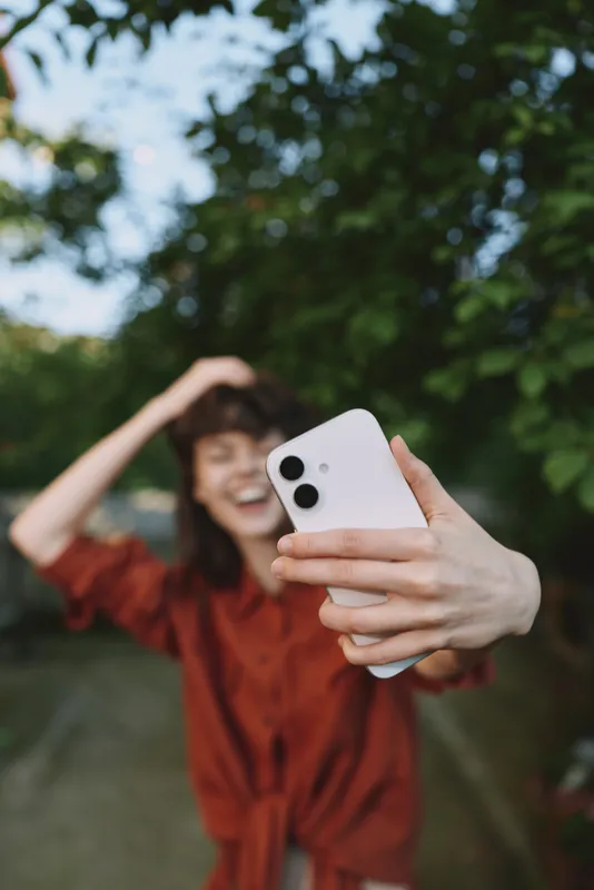 Smiling woman takes a selfie outdoors with a smartphone, wearing a red top. Bright natural light, greenery, and a blurred park background create a casual lifestyle portrait