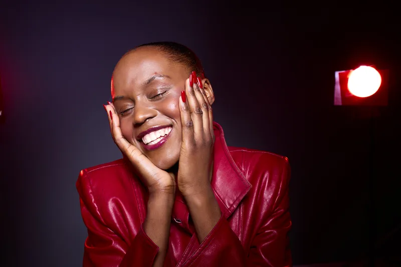 Happy black woman with dark skin wearing red leather jacket smiles with closed eyes and hands on face. Studio portrait with dark background and red spotlight. Concept of joy and confidence.