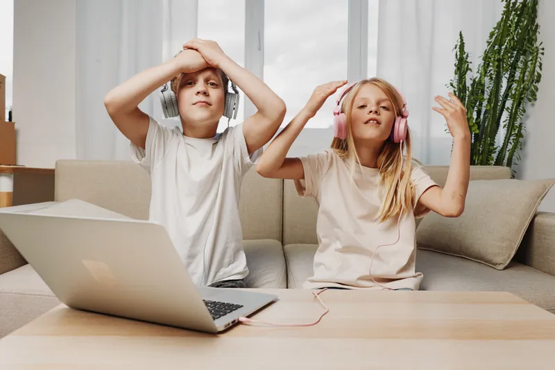 Two kids wearing headphones enjoy music while sitting on a couch, with a laptop on the table in a bright living room. Concept of digital entertainment, relaxation and childhood fun.