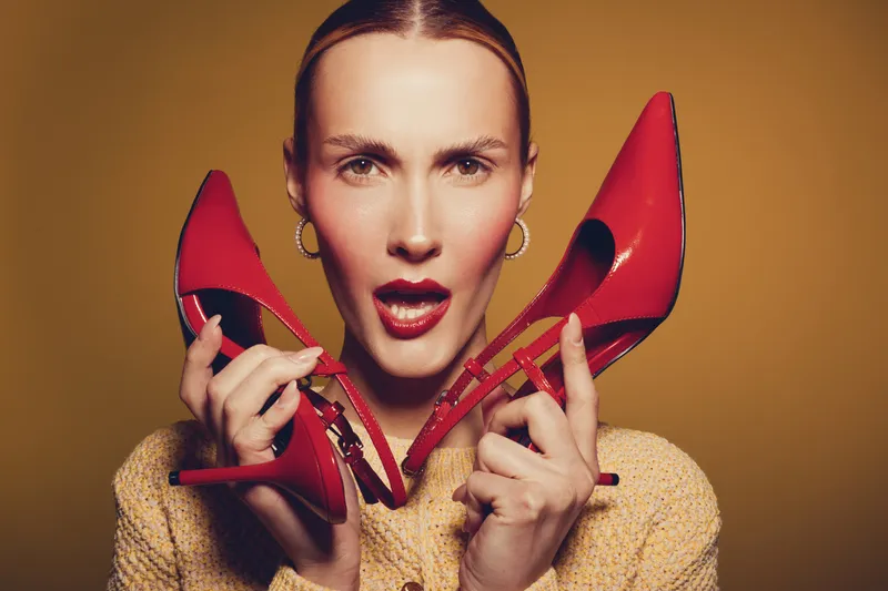 Young woman with red lipstick holds red high heels near face, wearing yellow textured jacket. Studio portrait on brown background. Concept of fashion, style, and beauty accessories.